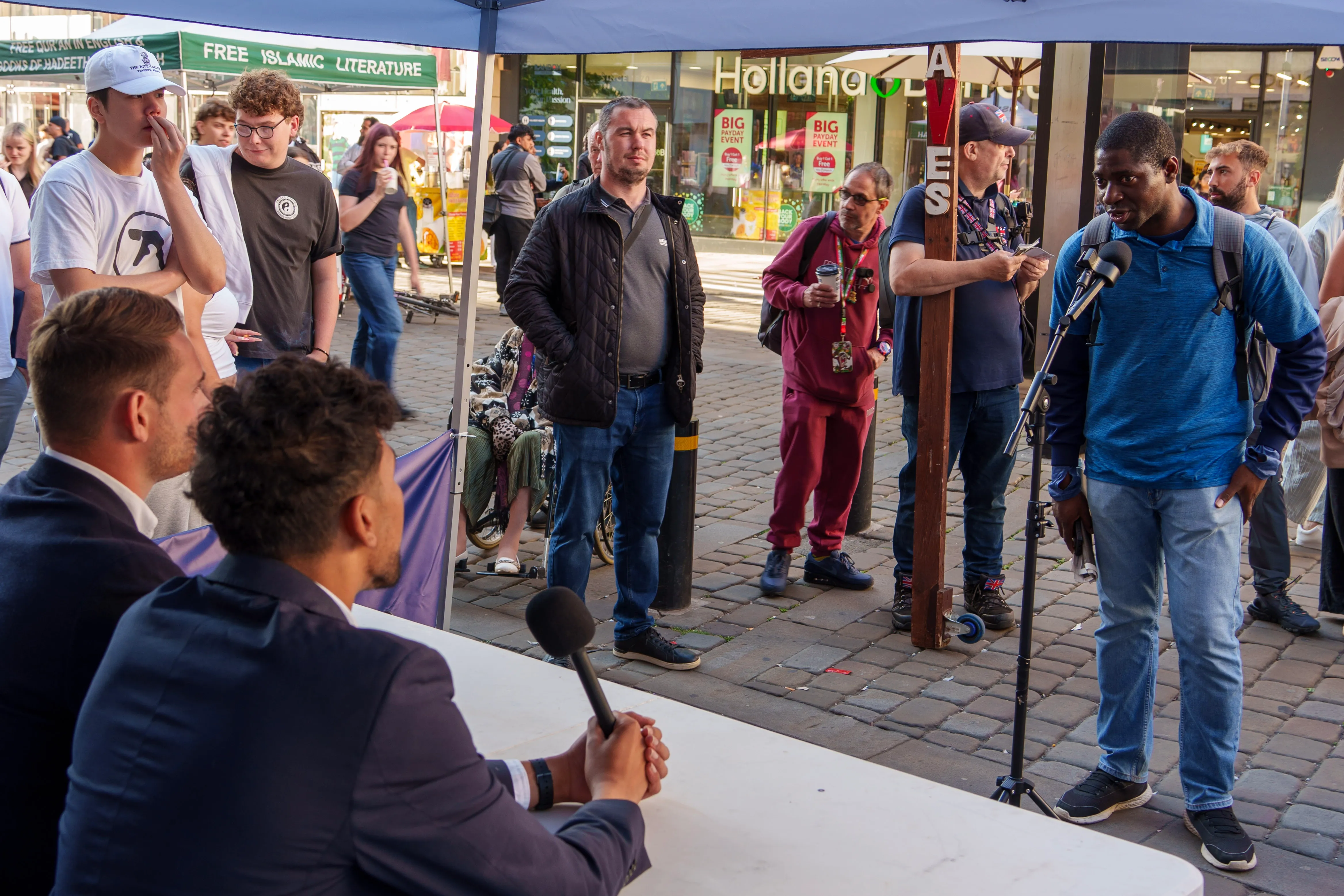 Public street debate in Manchester — a speaker at a mic while suited moderators listen from a table