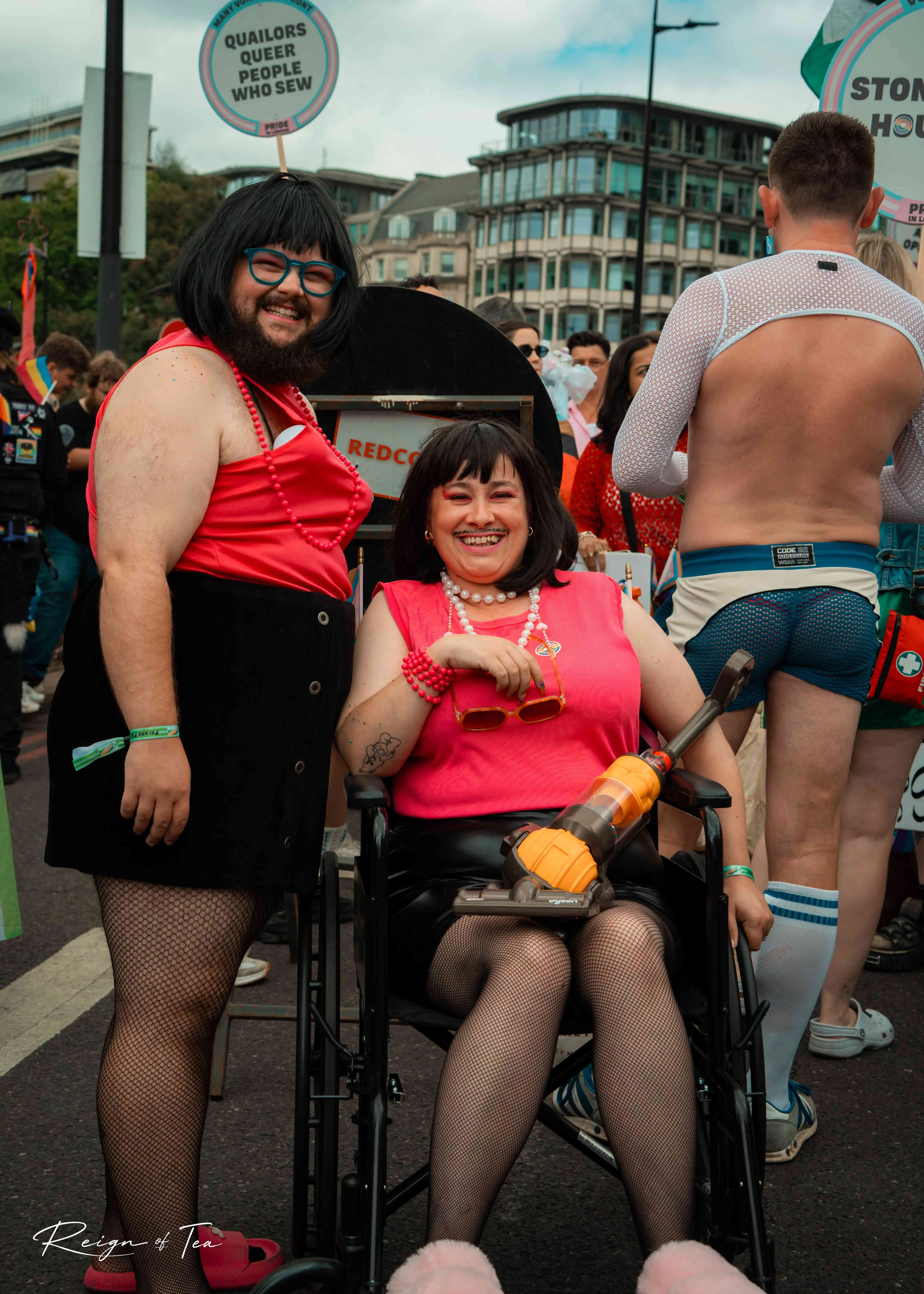 Two Pride parade participants in pink tops and black skirts, one seated with a toy vacuum cleaner